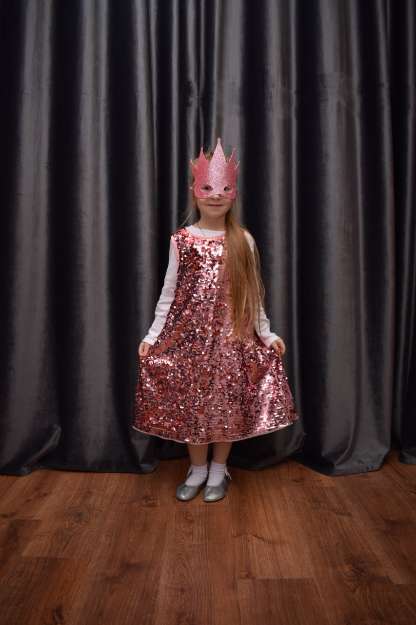 Dziewczynka w błyszczącej sukience i różowej koronie na tle szarej zasłony, prezentuje strój karnawałowy z lekkim uśmiechem.