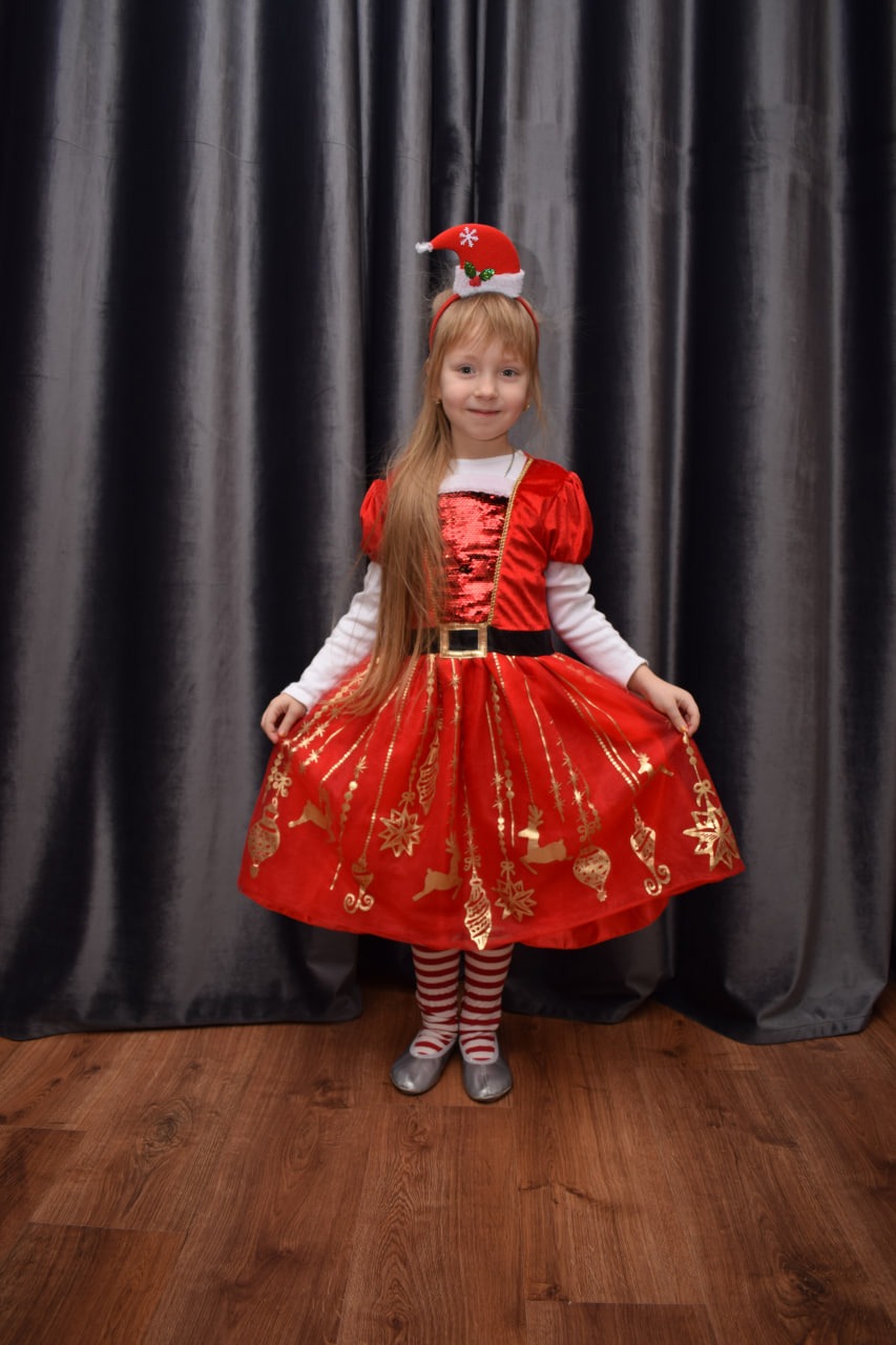 Dziewczynka w czerwonej sukience z motywami świątecznymi, w czapce Mikołaja z pomponem, pozuje na tle szarych zasłon i drewnianej podłogi.