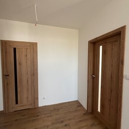 Konrad Rzemek Rzemass Bud - Nowe, drewniane drzwi wewnętrzne z przeszkleniami, zamontowane w świeżo wykończonym pokoju z panelami podłogowymi w odcieniach brązu, widoczny wystający kabel ze ściany pod sufitem.