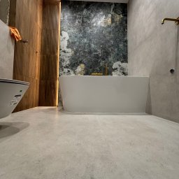 Konrad Rzemek Rzemass Bud - Nowoczesna łazienka po remoncie: wolnostojąca wanna na tle ściany z zielonego marmuru, drewniana boazeria, złota armatura i szara podłoga z płytek.