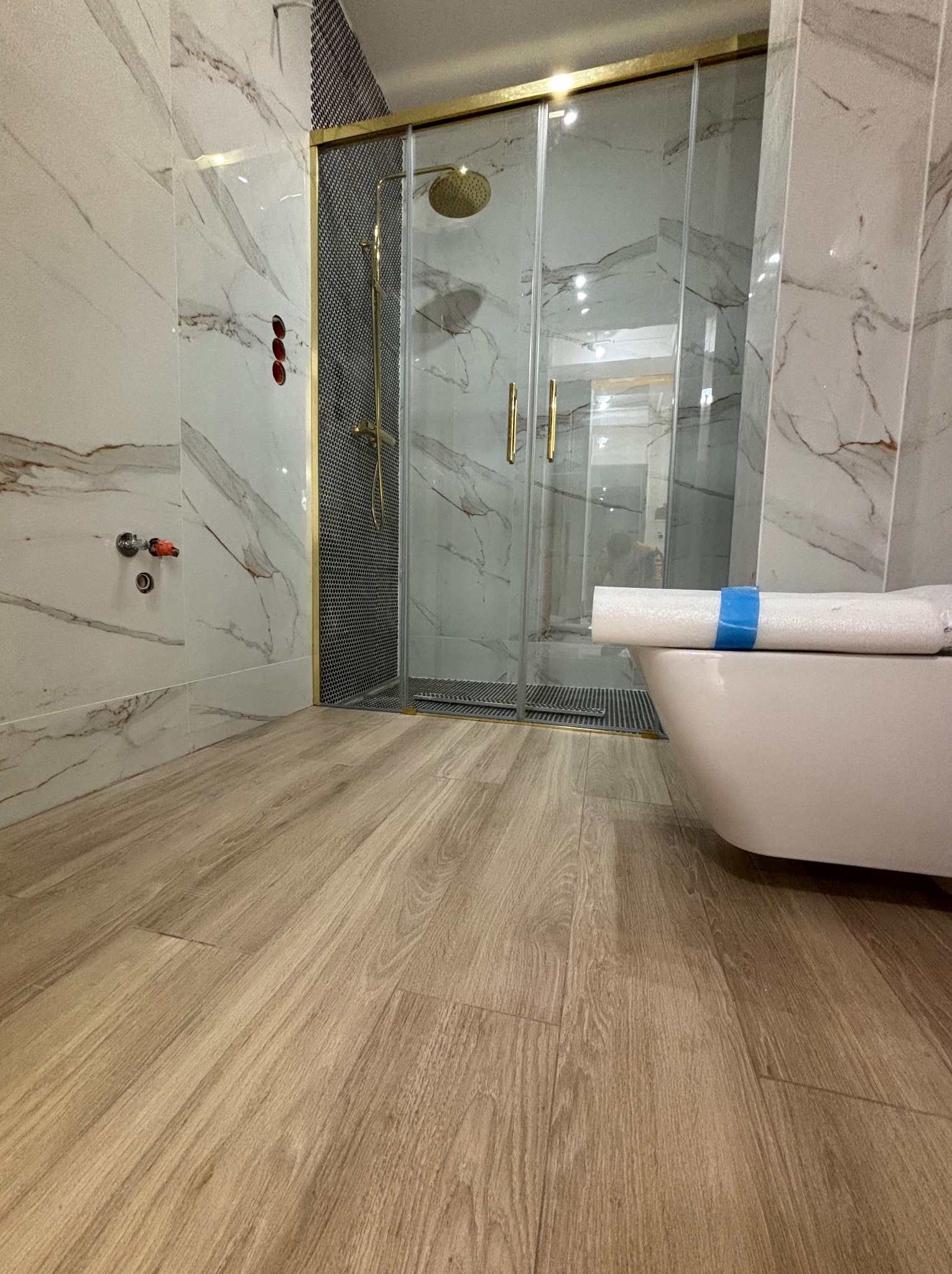 Nowoczesna łazienka w trakcie wykańczania, z marmurowymi płytkami na ścianach, drewnopodobną podłogą, kabiną prysznicową ze złotymi detalami i toaletą w folii ochronnej.