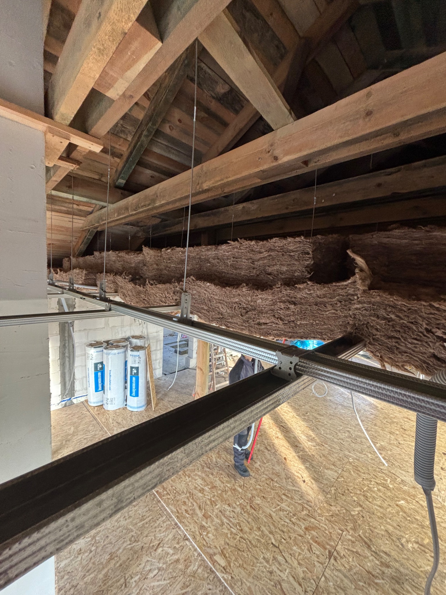 Montaż izolacji z wełny mineralnej pod drewnianą konstrukcją dachu na poddaszu, widoczne metalowe profile, rolki izolacji oraz fragment podłogi z płyt OSB.