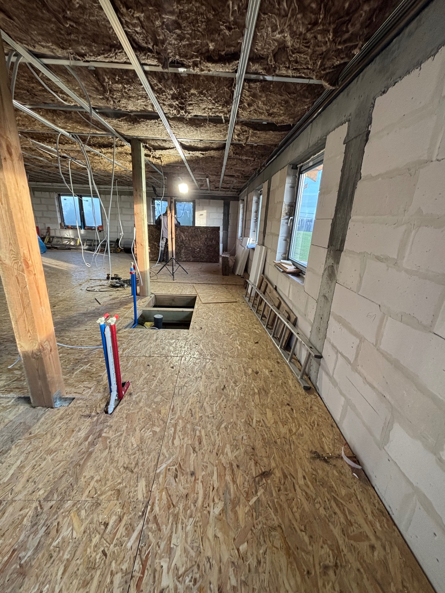 Wnętrze w trakcie prac wykończeniowych: surowa podłoga z płyt OSB, ściany z betonu komórkowego, izolacja wełną mineralną na suficie podwieszonym, rury instalacyjne pionowe z oznaczeniami...