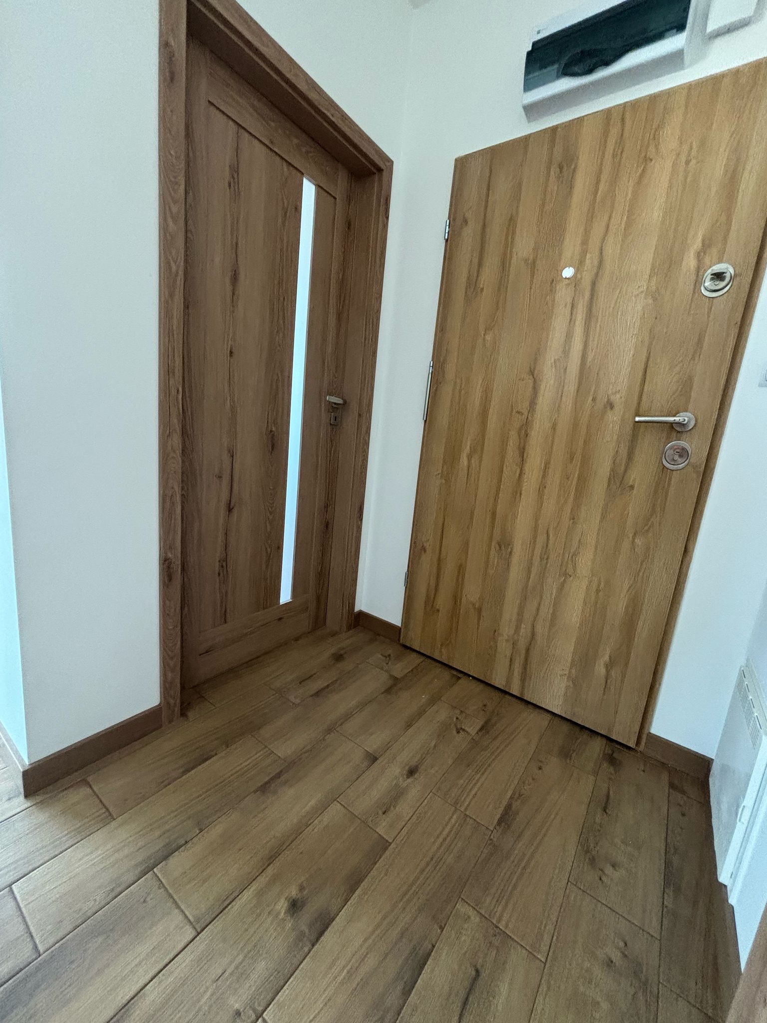 Dwa drewniane drzwi wewnętrzne o różnym designie, ułożone obok siebie na tle drewnopodobnych płytek podłogowych i białych ścian.