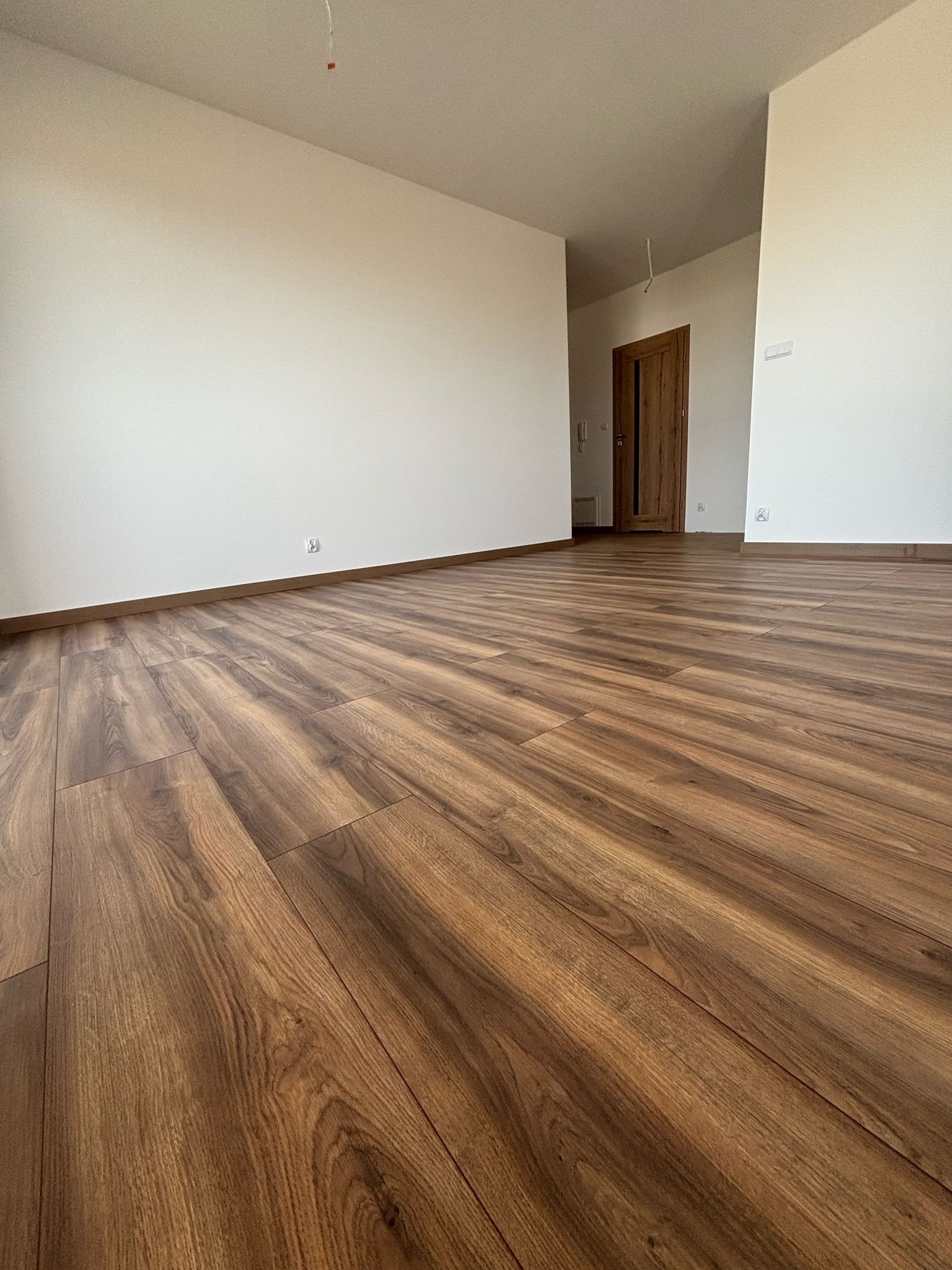 Nowo ułożone panele podłogowe w pustym pokoju z białymi ścianami, widoczne listwy przypodłogowe i drewniane drzwi w tle.