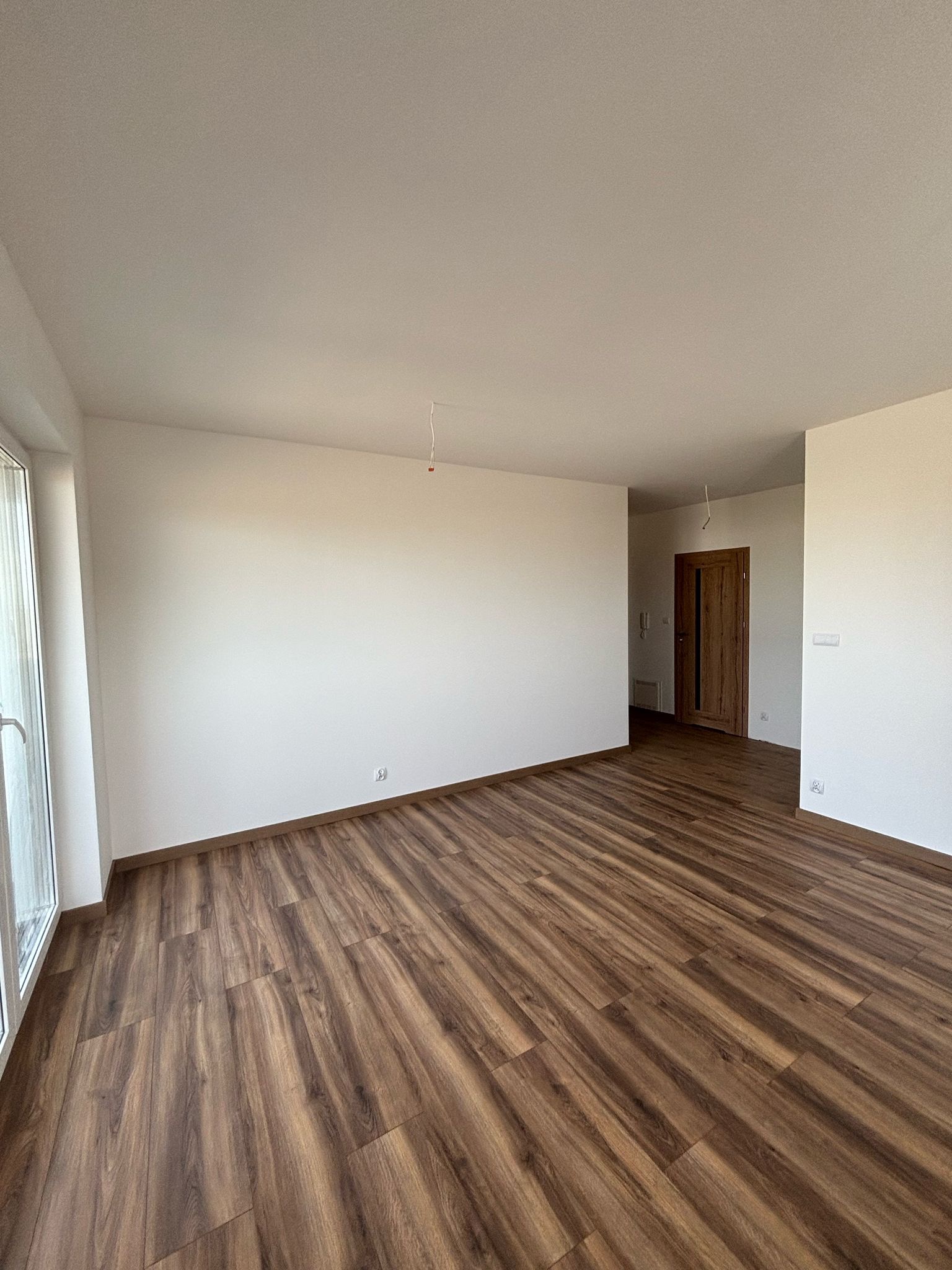 Nowe panele podłogowe w odcieniach brązu, świeżo położone w pustym pokoju z białymi ścianami i oknem po lewej stronie, widoczne gniazdka elektryczne, brak listew przypodłogowych.