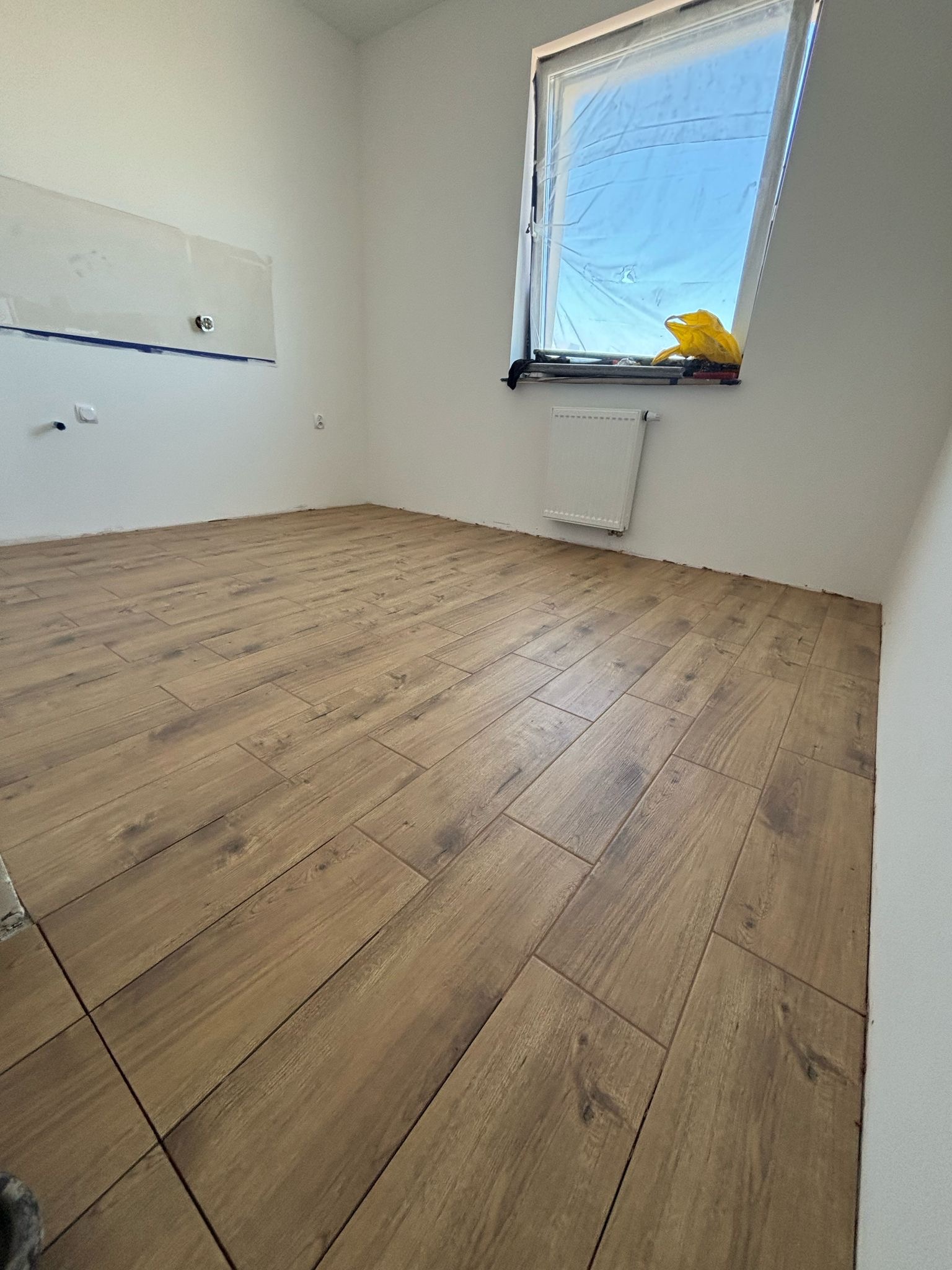 Ułożone płytki podłogowe imitujące drewno w jasnym pomieszczeniu z oknem zabezpieczonym folią malarską i grzejnikiem.