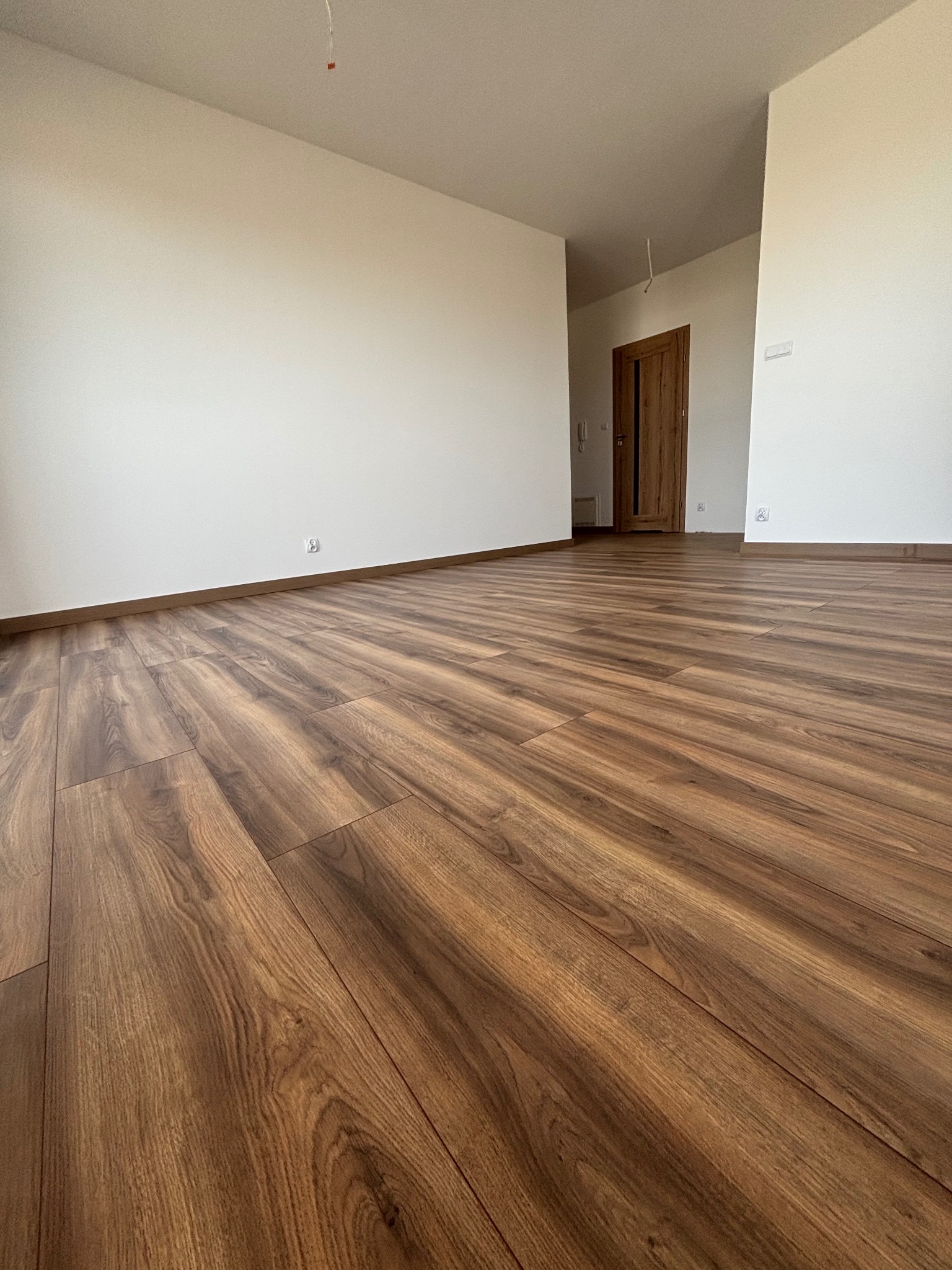 Nowo ułożone, brązowe panele podłogowe w pustym pokoju z białymi ścianami i drewnianymi listwami przypodłogowymi, widoczny fragment drzwi w tle.