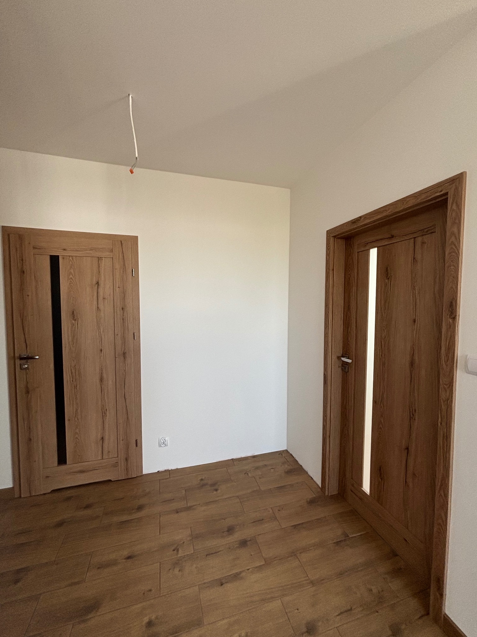 Nowe, drewniane drzwi wewnętrzne z przeszkleniami, zamontowane w świeżo wykończonym pokoju z panelami podłogowymi w odcieniach brązu, widoczny wystający kabel ze ściany pod sufitem.
