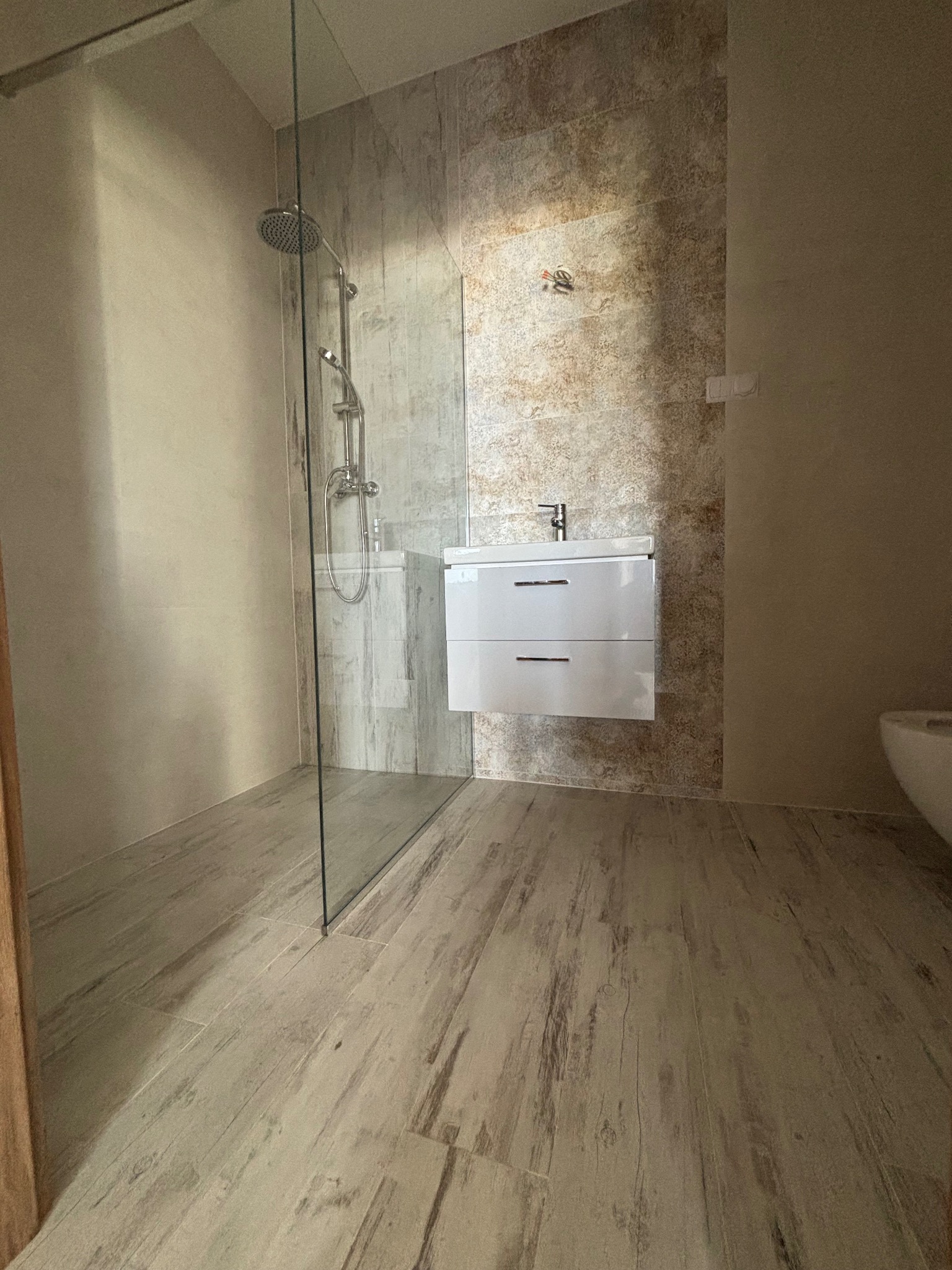 Nowoczesna łazienka z kabiną prysznicową typu walk-in, umywalką z szafką w kolorze białym i płytkami imitującymi drewno na podłodze, z widocznymi drobnymi niedociągnięciami w wykończeniu.