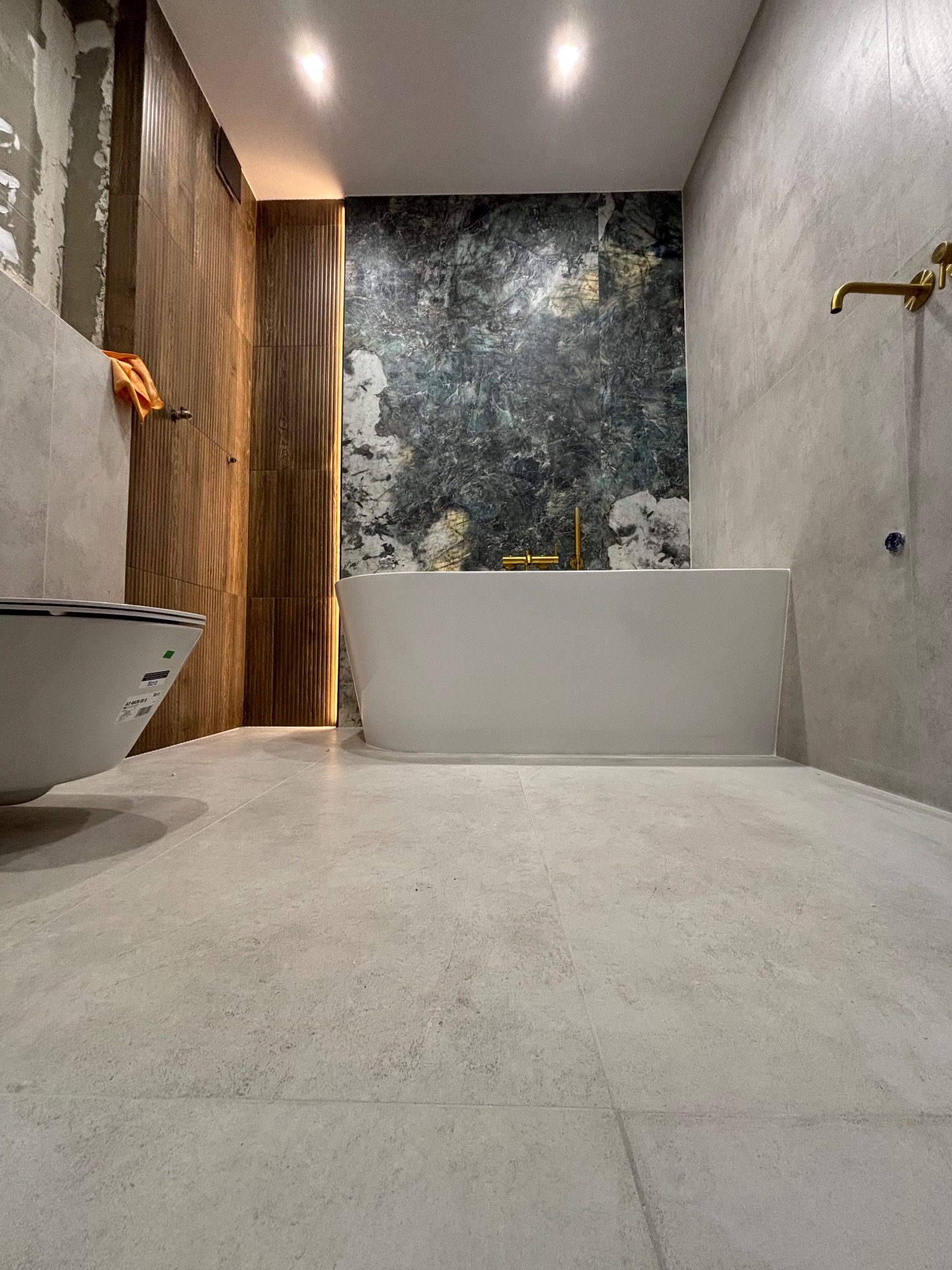 Nowoczesna łazienka z wanną, marmurową ścianą i drewnianymi panelami. Widoczne złote elementy armatury i szara podłoga. Częściowo widoczna toaleta.