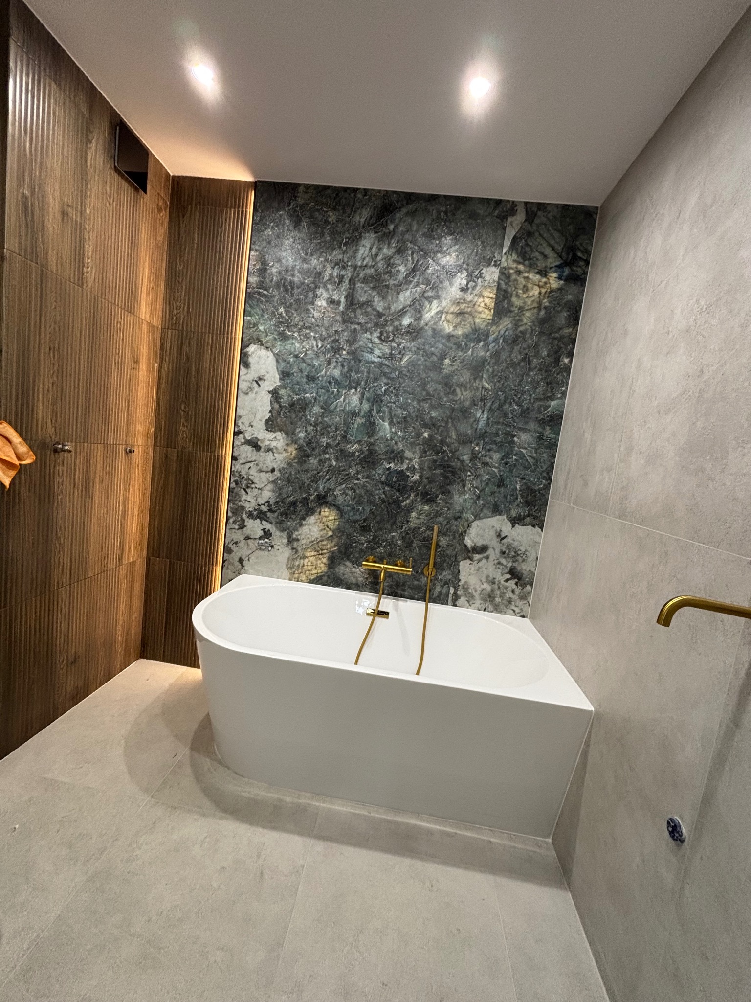 Nowoczesna łazienka z wolnostojącą wanną, drewnianą okładziną ściany z subtelnym oświetleniem i marmurową ścianą. Złote akcenty baterii i armatury.