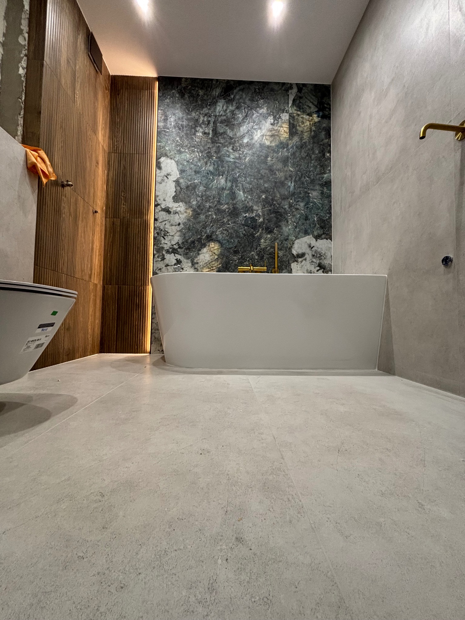 Nowoczesna łazienka po remoncie: wolnostojąca wanna na tle ściany z zielonego marmuru, drewniana boazeria, złota armatura i szara podłoga z płytek.