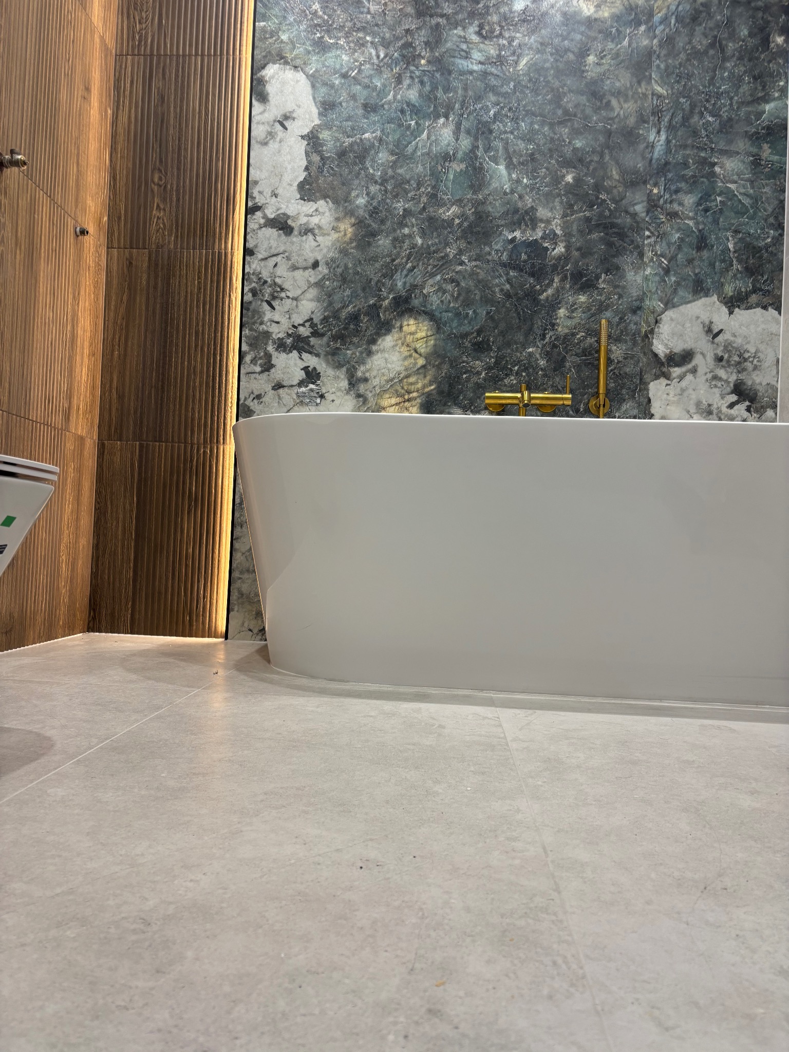 Nowoczesna łazienka z wolnostojącą wanną na tle ściany z płytek imitujących marmur i drewnianych paneli. Złota bateria wannowa.