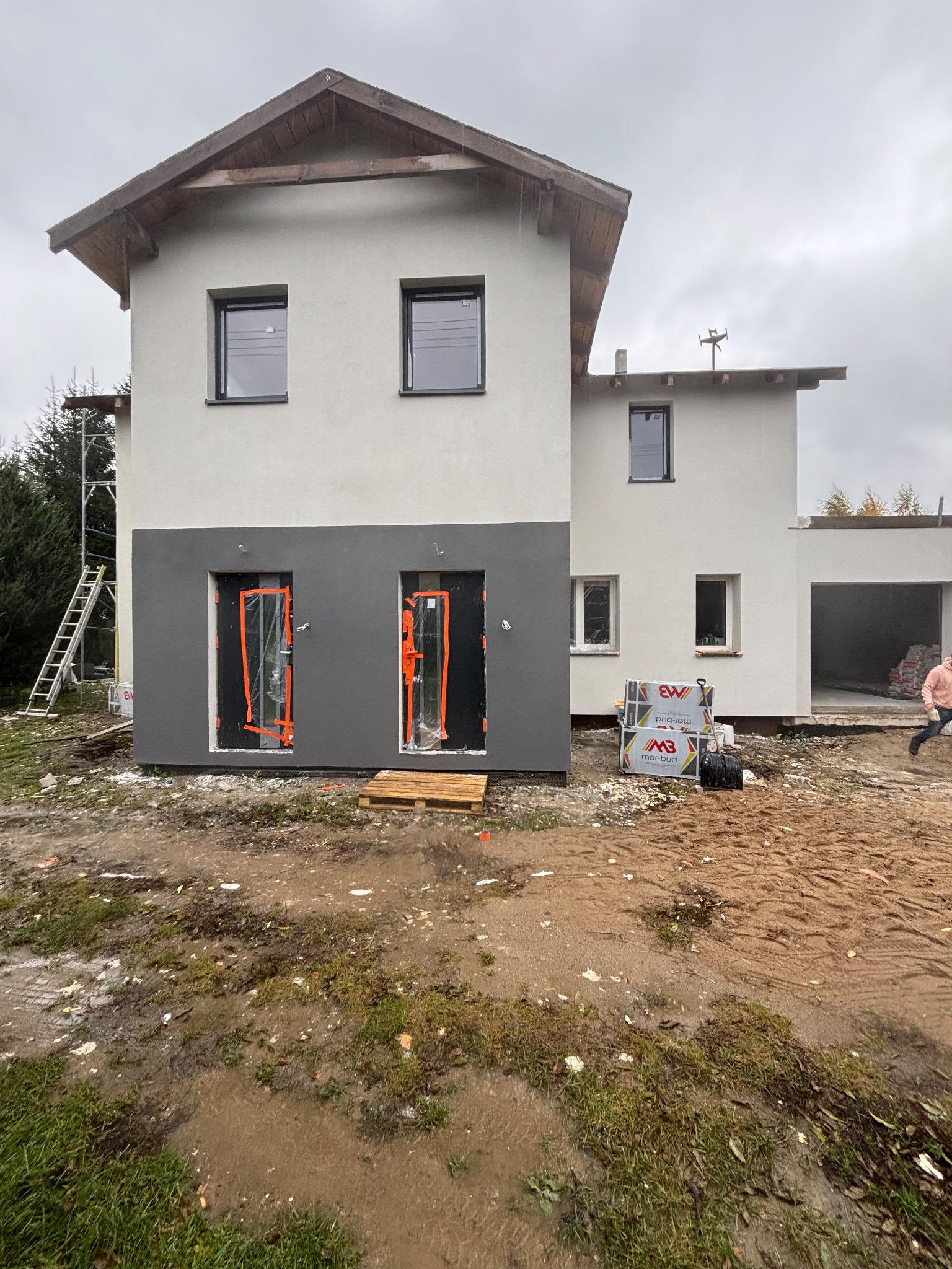 Dwukondygnacyjny dom z szarą dolną elewacją i zabezpieczonymi folią drzwiami wejściowymi. Na zewnątrz widoczne materiały budowlane i drabina.