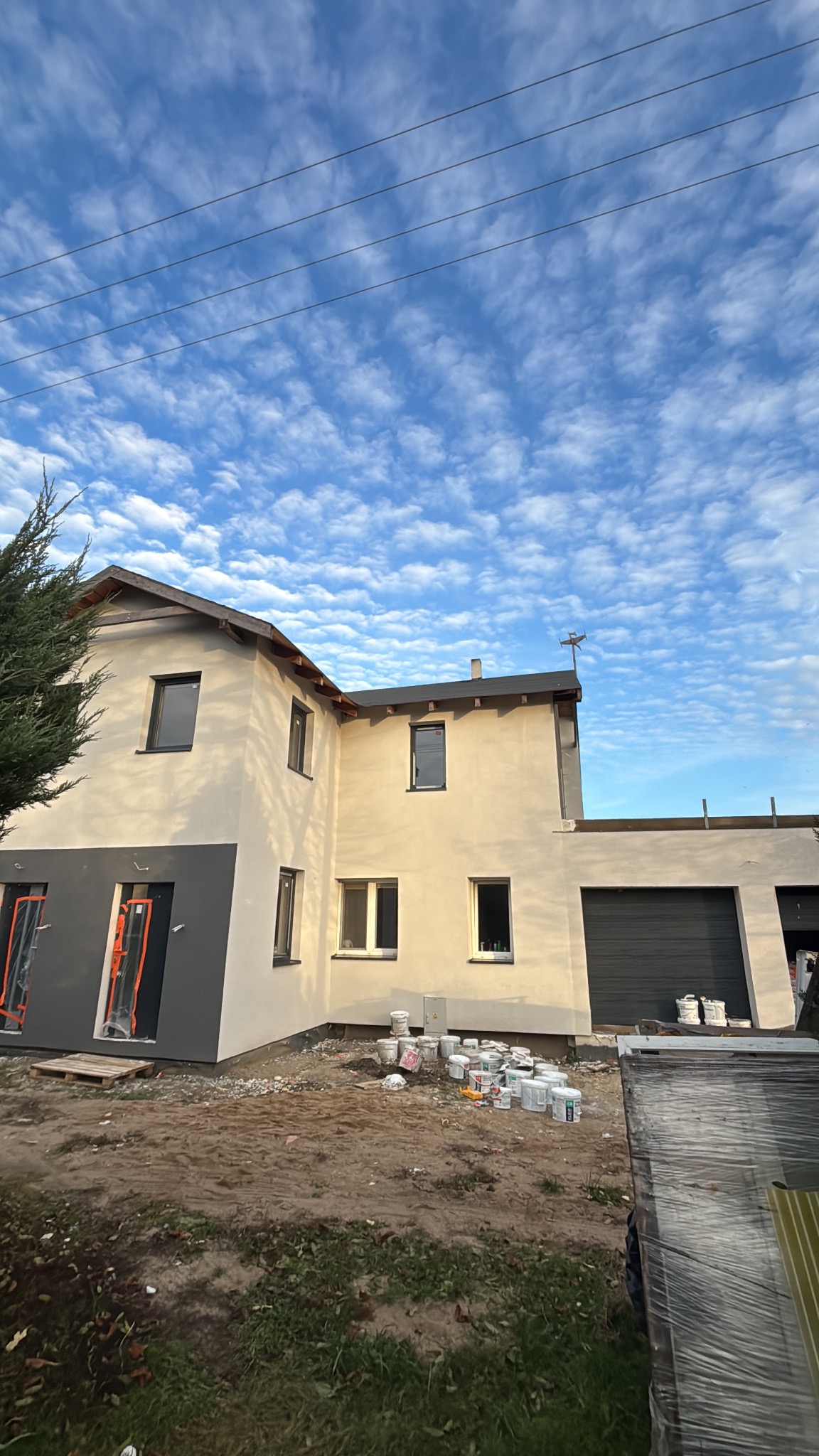 Dwukondygnacyjny dom z garażem w trakcie ocieplania elewacji, widoczne puszki z farbą i zabezpieczone folią okna. Błękitne niebo z obłokami w tle.