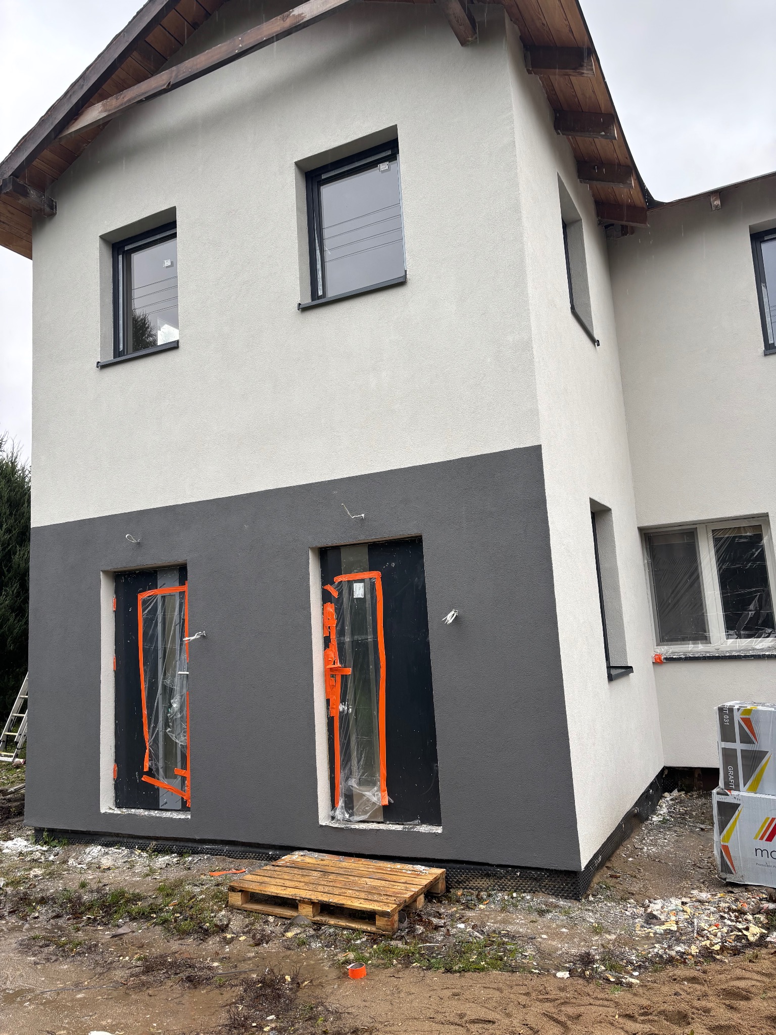 Dwukolorowa elewacja domu jednorodzinnego w trakcie prac wykończeniowych. Okna i drzwi zabezpieczone folią. Paleta i materiały budowlane obok budynku.