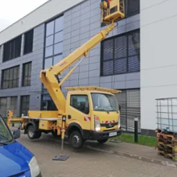 Żółty podnośnik koszowy na tle biurowca z szarymi panelami, pracownik w koszu myje okna z żaluzjami, widoczny zbiornik na wodę na palecie.