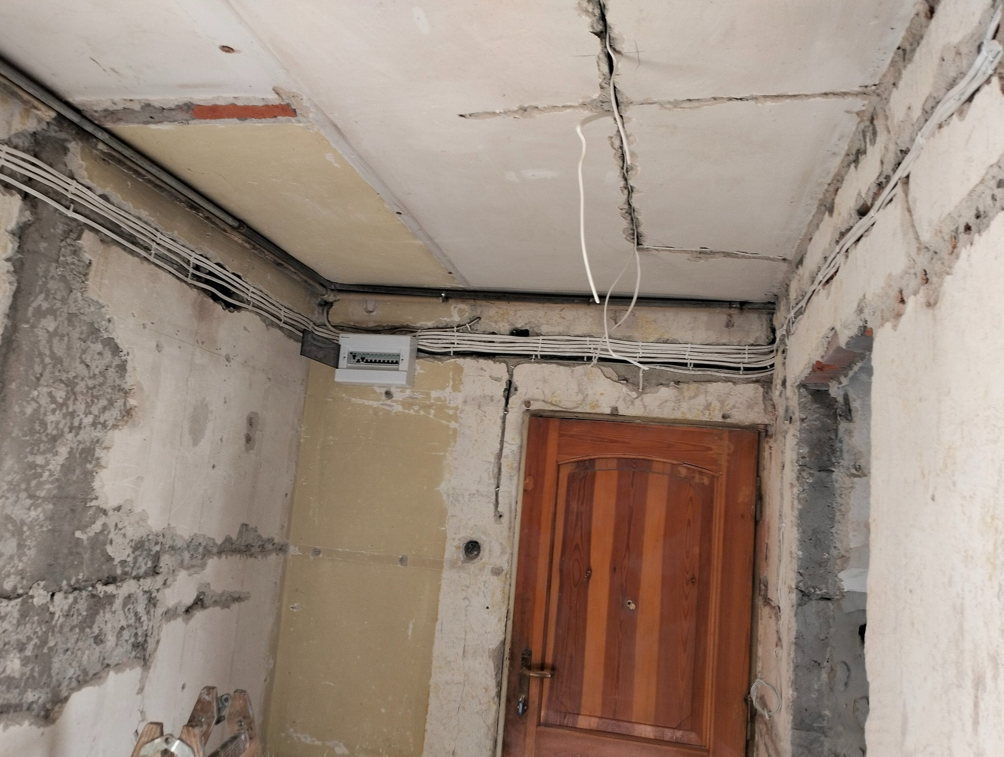 Wnętrze w trakcie remontu z widoczną instalacją elektryczną: kable prowadzone po suficie i ścianach, tablica rozdzielcza na ścianie, surowy tynk i fragment drewnianych drzwi.