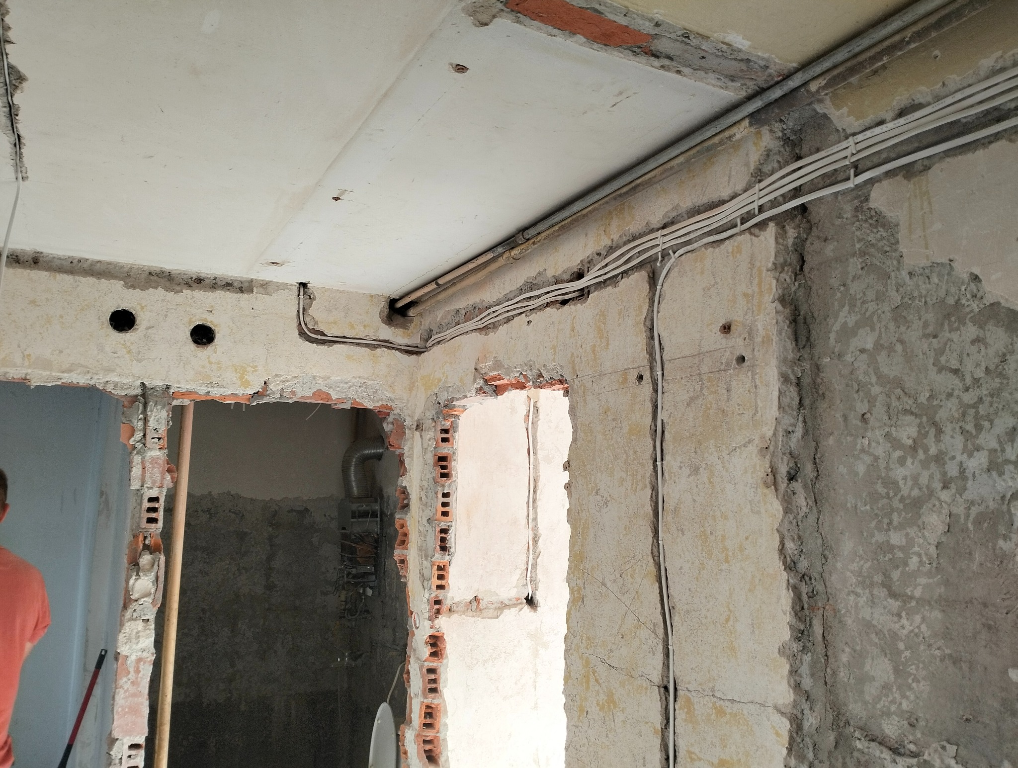 Wnętrze w trakcie remontu z odsłoniętymi przewodami elektrycznymi biegnącymi wzdłuż sufitu i ścian, widoczne fragmenty cegieł i instalacji gazowej w sąsiednim pomieszczeniu.