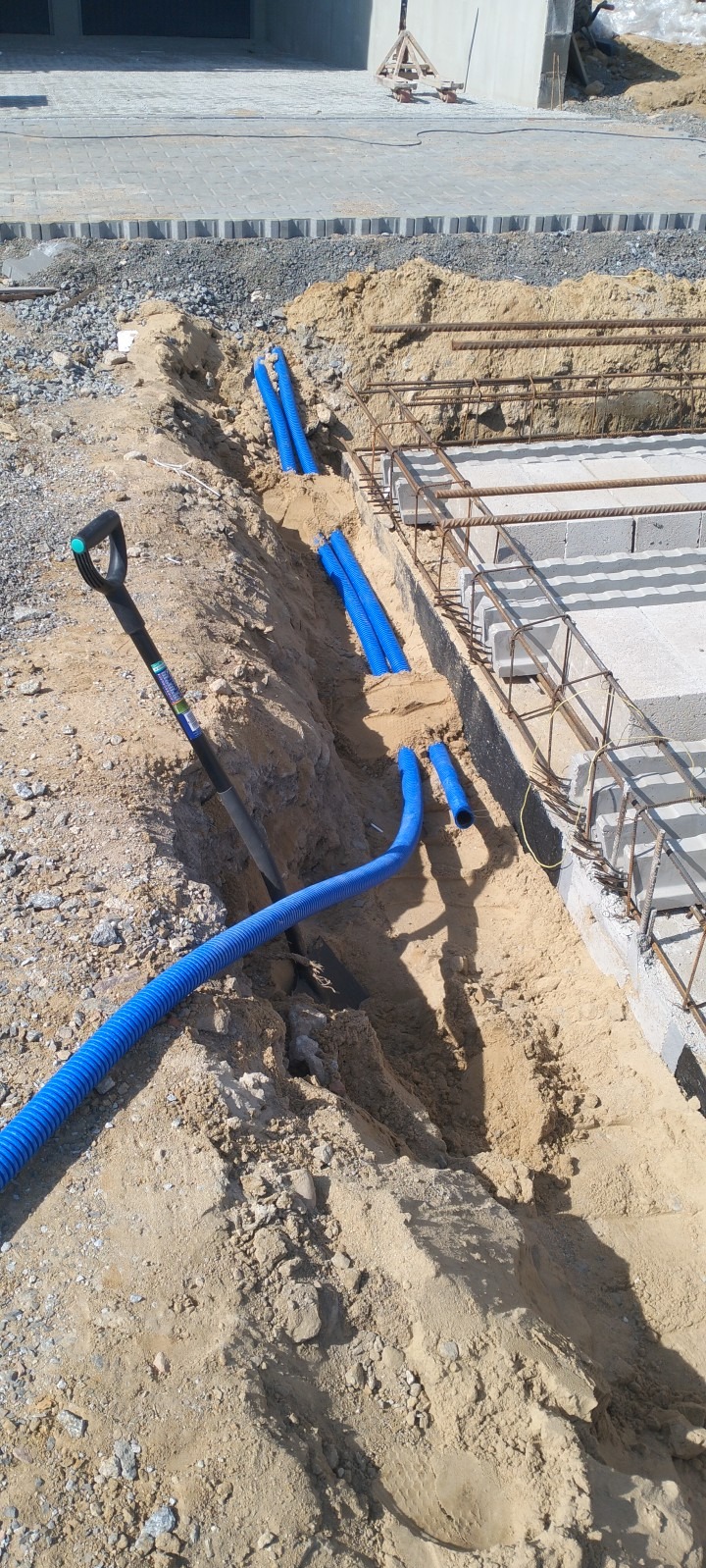 Wykop z niebieskimi rurami karbowanymi ułożonymi w ziemi, obok fundamentów budynku w trakcie budowy, widoczna łopata wbita w ziemię.