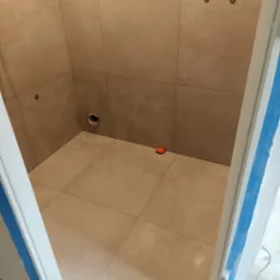 Wykafelkowana kabina prysznicowa z beżowymi, kwadratowymi płytkami, widoczne otwory na armaturę i pomarańczowy krzyżyk dystansowy, maskująca taśma malarska na ościeżnicy.