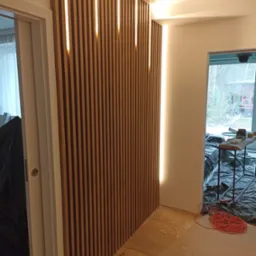 Drewniana ścianka działowa z pionowych listew z wbudowanym oświetleniem LED, widok na częściowo zakryte pomieszczenie w trakcie prac wykończeniowych.