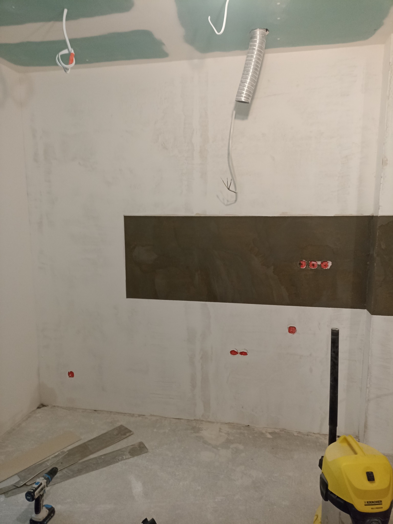 Surowa ściana w trakcie wykańczania z widocznymi przewodami elektrycznymi, fragmentem płyty karton-gips na suficie, w tle odkurzacz przemysłowy i narzędzia.