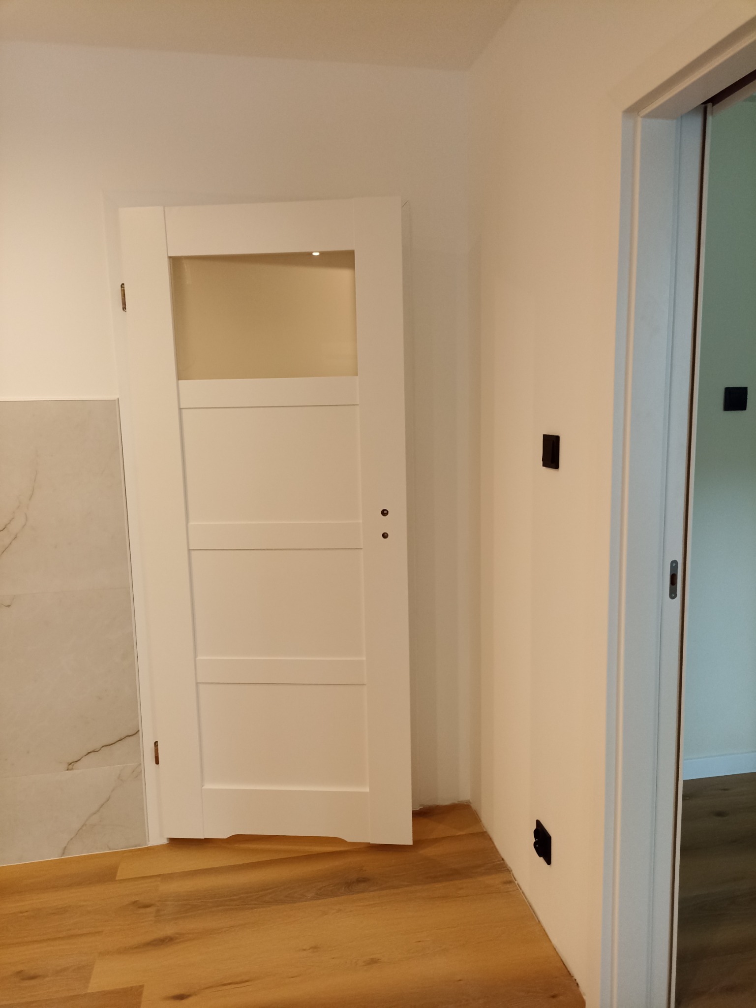 Białe drzwi z szybą w nowo wyremontowanym wnętrzu, widoczne płytki imitujące marmur i drewniana podłoga.