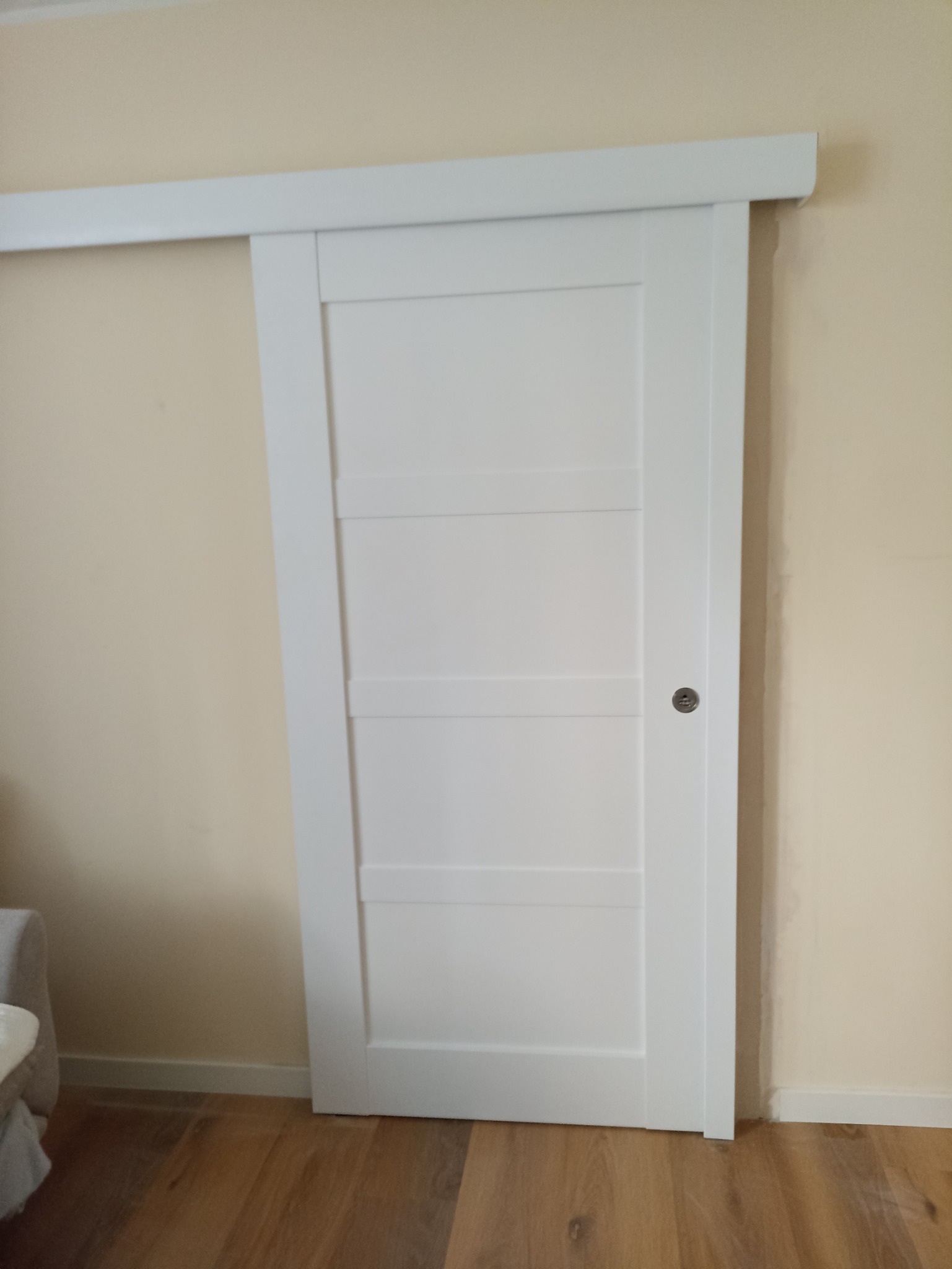 Białe, przesuwne drzwi wewnętrzne z czterema panelami, zamontowane na szynie w jasnym wnętrzu z drewnianą podłogą.