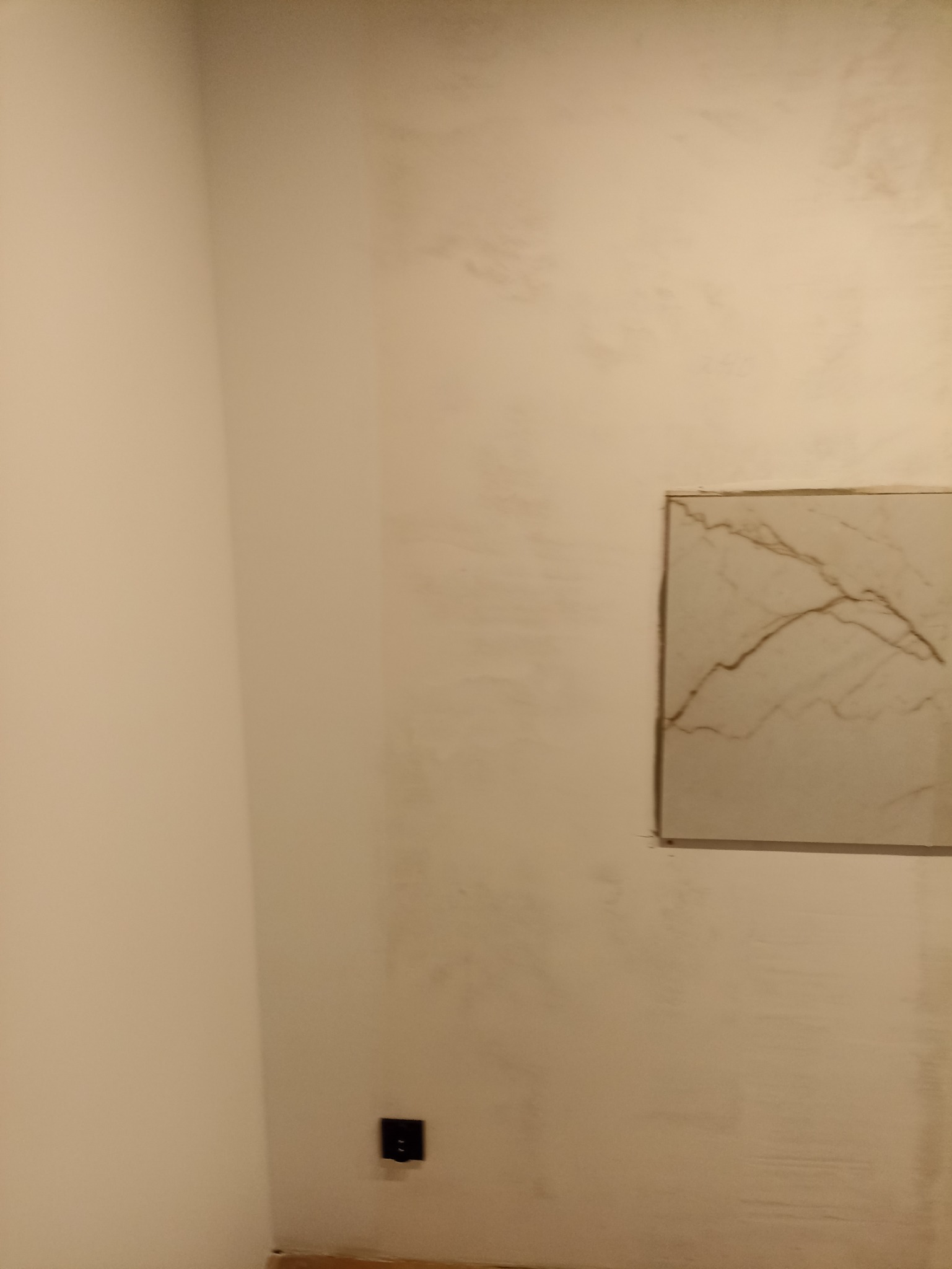 Fragment ściany w trakcie prac wykończeniowych z wstawionym kwadratowym elementem imitującym marmur, widoczne ślady po szpachlowaniu i gniazdko elektryczne.