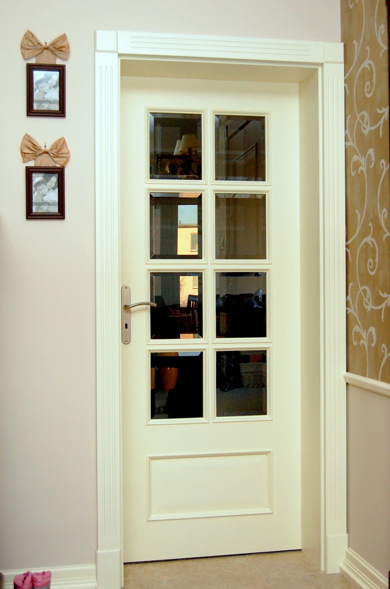 Białe drzwi z ośmioma szklanymi szybkami, widoczne odbicia wnętrza pokoju, na ścianie dekoracje i zdjęcia w ramkach.