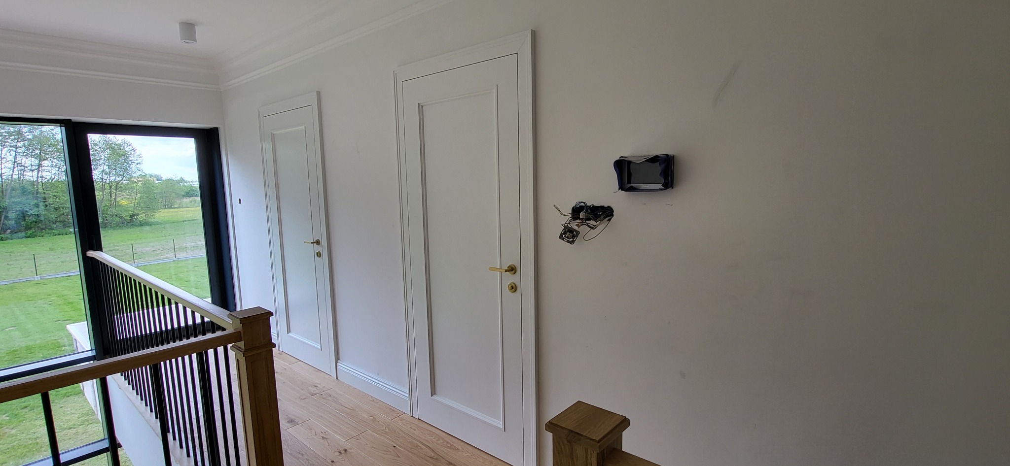 Korytarz z dwiema białymi skrzydłami drzwiowymi z widocznymi złotymi klamkami, drewnianą podłogą, balustradą schodów i widokiem na zieleń za oknem. Na ścianie widoczna instalacja elektryczna...