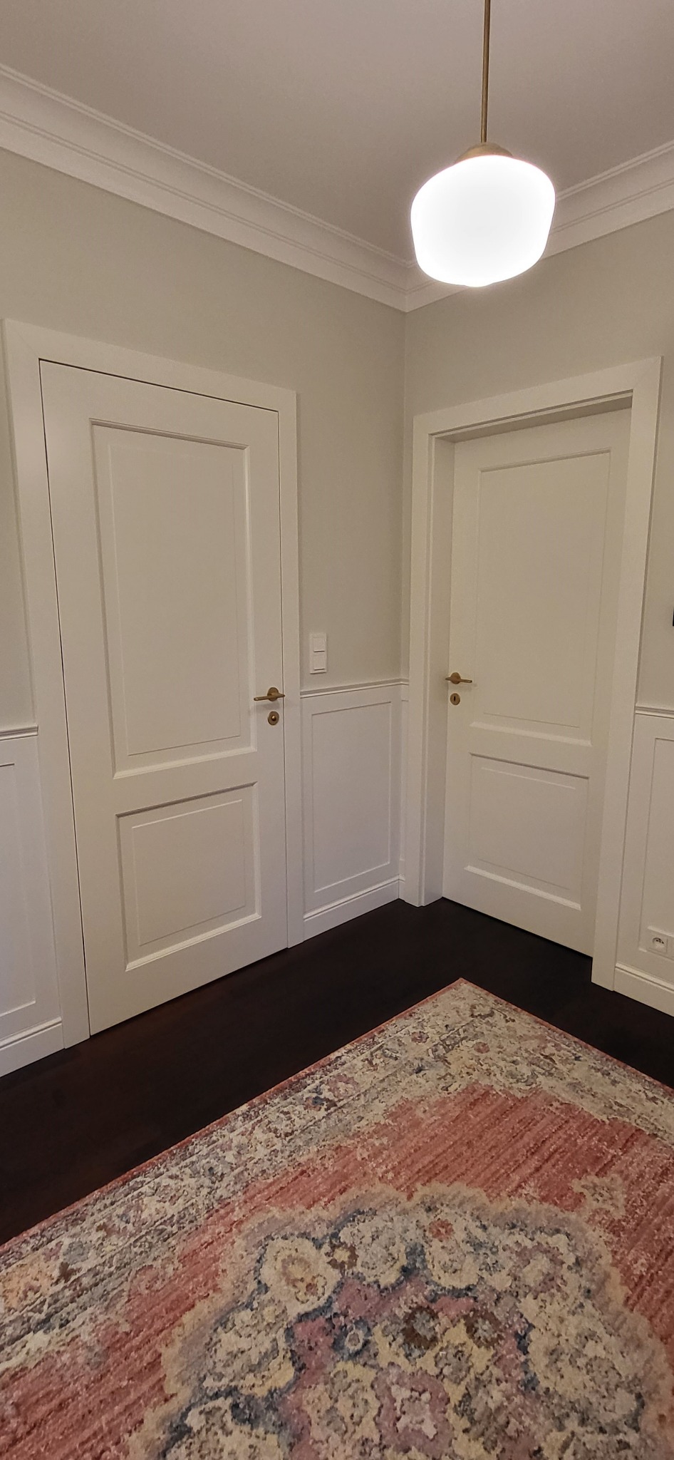 Dwie białe, panelowe konstrukcje z mosiężnymi klamkami w korytarzu z dekoracyjnymi panelami ściennymi i wzorzystym dywanem w odcieniach różu i szarości.