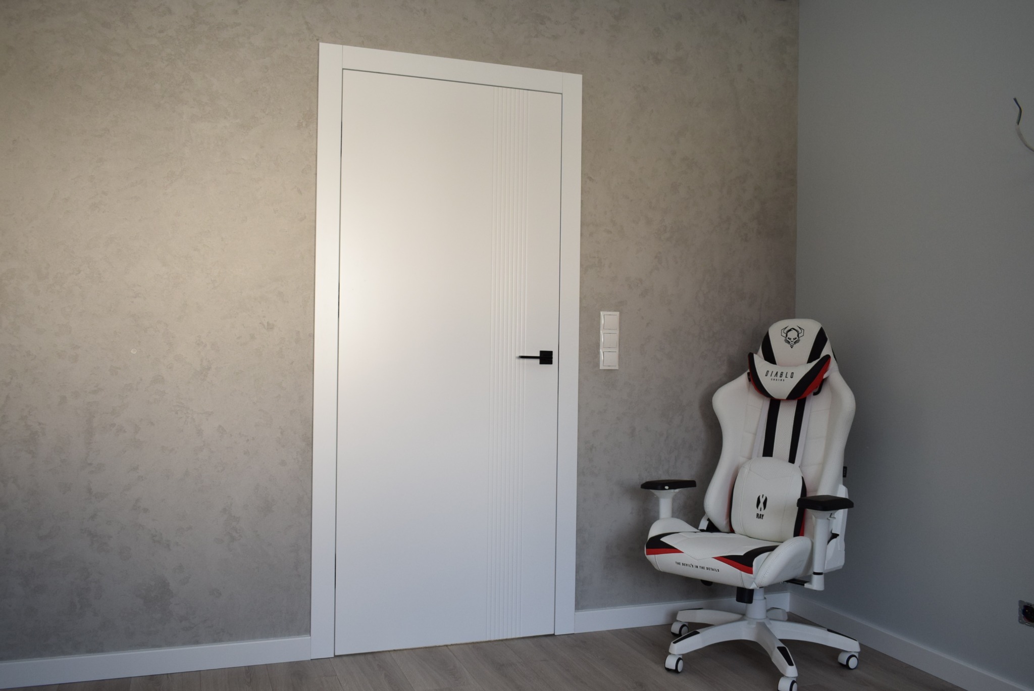 Białe drzwi wewnętrzne z czarną klamką i pionowymi frezowaniami, widoczne na tle ściany o fakturze betonu, obok stoi gamingowe krzesło w kolorze białym.