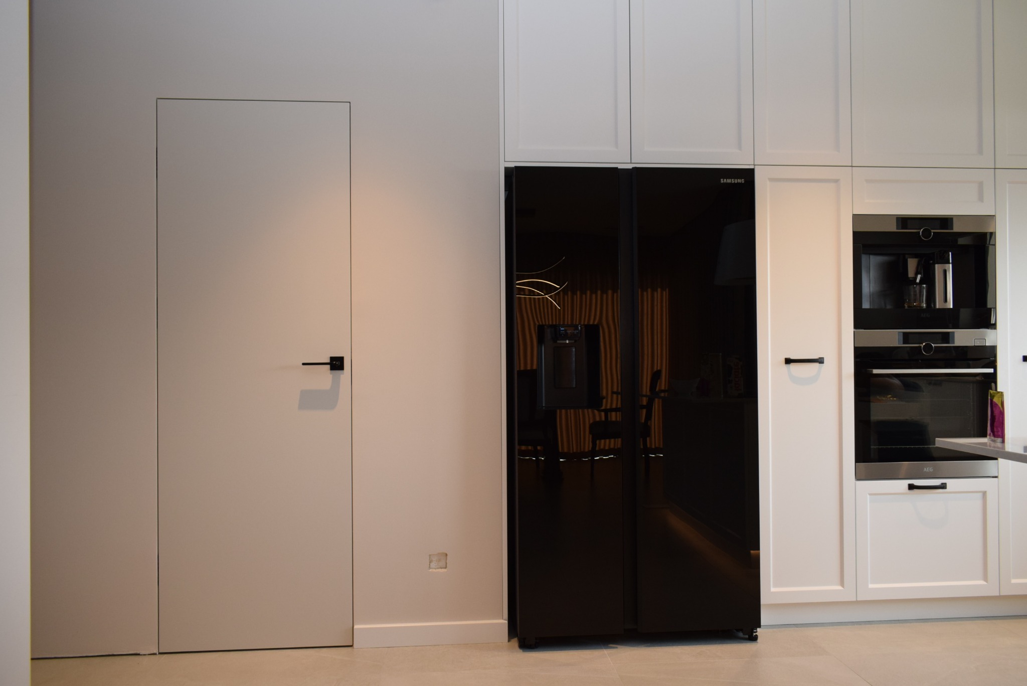 Minimalistyczna kuchnia z ukrytymi drzwiami w kolorze śmietanki, z czarną klamką, sąsiadującymi z zabudową kuchenną i czarną lodówką Samsung.