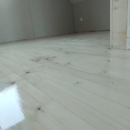 PlenerMen - Jasne, lakierowane deski podłogowe w nowym poddaszu z płytami karton-gips na ścianach i drewnianą konstrukcją dachu. Widok z dołu, blisko podłogi.