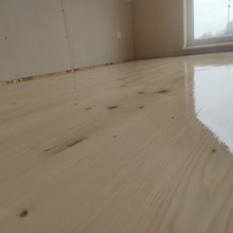 PlenerMen - Jasne, lakierowane panele podłogowe z widocznymi sękami, świeżo położone przy ścianie z płyt karton-gips i oknem w tle, ukazujące gładką, błyszczącą powierzchnię.
