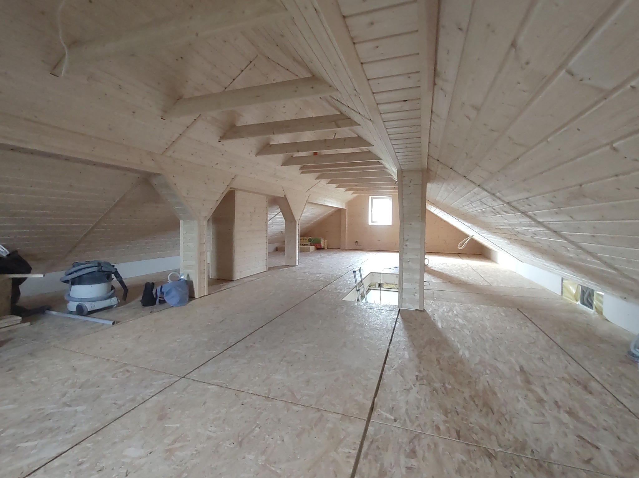 Jasne drewniane wnętrze poddasza z widoczną konstrukcją szkieletową, deskami podłogowymi i oknem w tle. Częściowo widoczny odkurzacz.