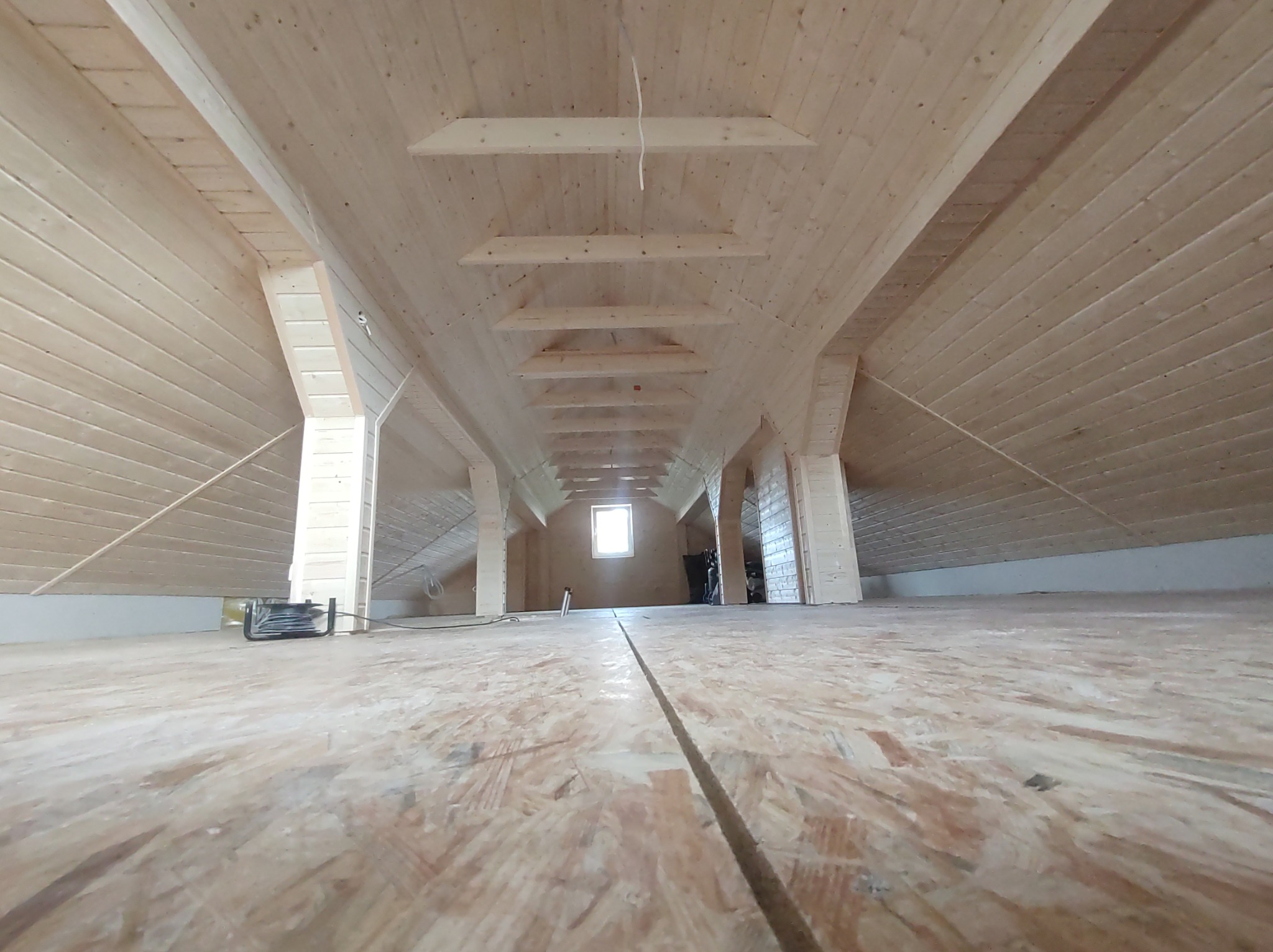 Surowe drewno na poddaszu z widocznymi belkami stropowymi i oknem w oddali. Podłoga z płyt OSB. Widok z niskiej perspektywy.