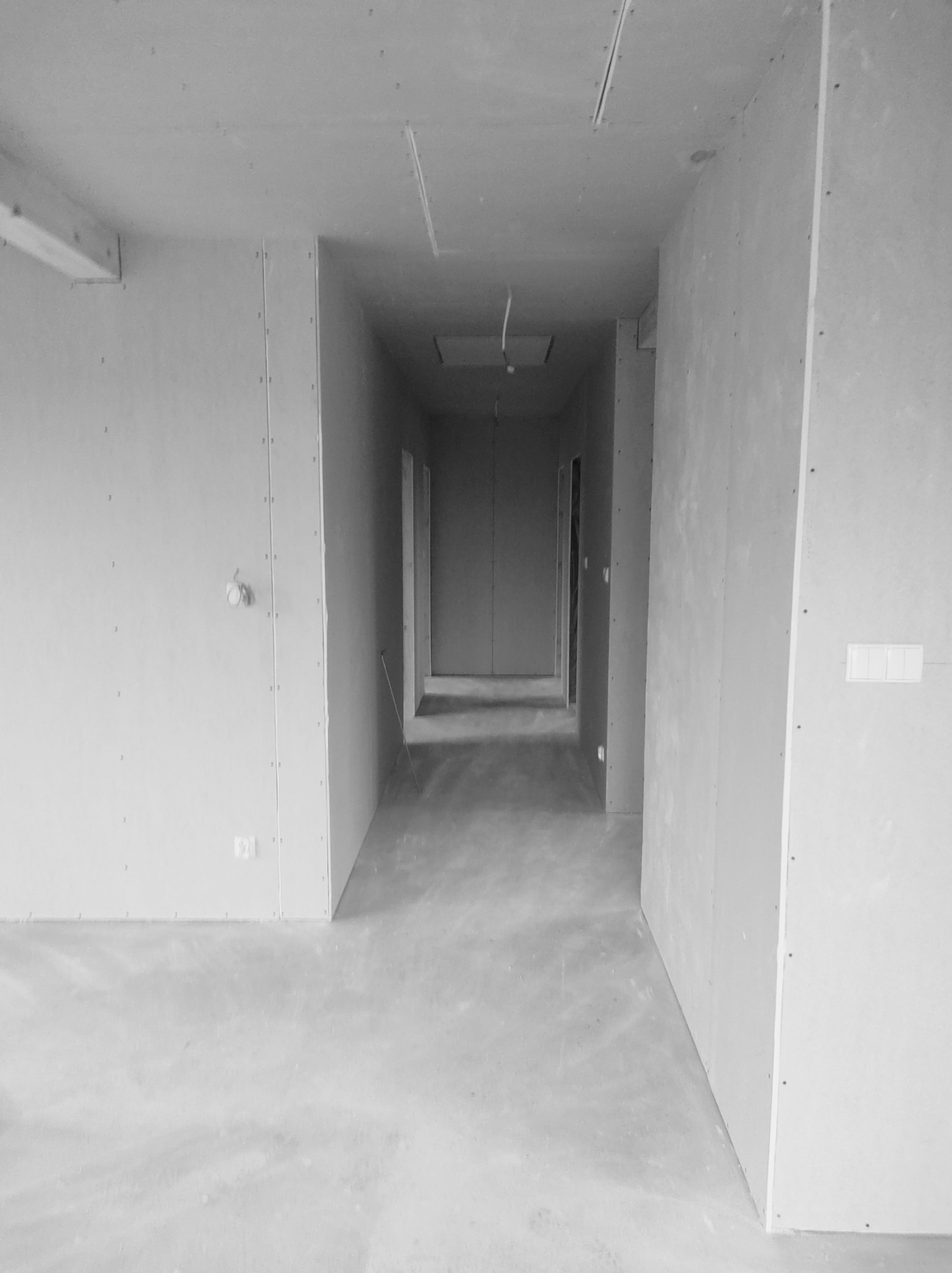 Wnętrze w trakcie prac wykończeniowych: ściany obłożone płytami gipsowo-kartonowymi, surowy beton na podłodze, widok na korytarz z kilkoma otworami drzwiowymi.