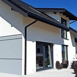 KRASBUD - Nowoczesny dom jednorodzinny z białymi ścianami, czarną stolarką okienną i dachem, z elementami szarego wykończenia elewacji oraz meblami ogrodowymi na tarasie.