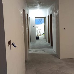 KRASBUD - Korytarz w trakcie prac wykończeniowych, ściany pokryte świeżo nałożoną gładzią szpachlową, widoczne ślady po szpachli, przygotowane pod malowanie, w tle drabina i otwarte drzwi.