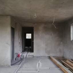 KRASBUD - Surowe ściany w trakcie prac wykończeniowych, widoczne kable elektryczne zwisające z sufitu oraz ułożone na podłodze rury instalacyjne i deski.