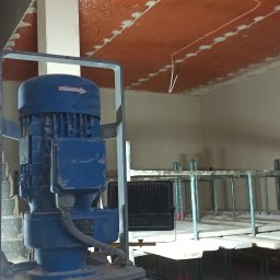 KRASBUD - Widok na maszynę do tynkowania Kaleta, z surowym, pomarańczowym sufitem w tle, gdzie widoczne są ślady szpachlowania połączeń płyt.