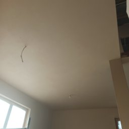 KRASBUD - Wnętrze w trakcie prac wykończeniowych, widoczny sufit po nałożeniu gładzi, z wystającym przewodem elektrycznym, oraz fragment ściany. W tle widoczne okno i rusztowanie.