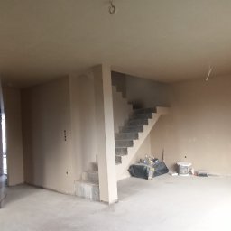 KRASBUD - Wnętrze domu w Iławie po tynkowaniu, widoczne schody, ściany w jasnym odcieniu, narzędzia pracy i pozostałości po pracach wykończeniowych.