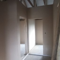 KRASBUD - Surowe wnętrze poddasza z nałożonymi tynkami na ścianach, widoczne drewniane elementy konstrukcji dachu i przygotowane otwory drzwiowe.