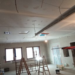 KRASBUD - Wnętrze w trakcie remontu: sufit i ściany po szpachlowaniu, widoczne drabiny, rusztowanie, kable zwisające z sufitu, okna i zabezpieczone folią drzwi.