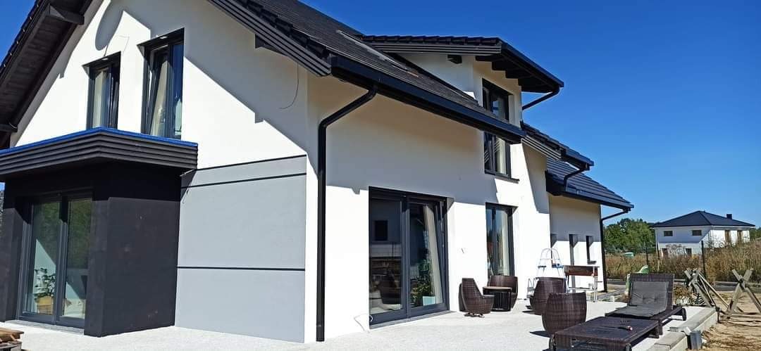 Nowoczesny dom jednorodzinny z białymi ścianami, czarną stolarką okienną i dachem, z elementami szarego wykończenia elewacji oraz meblami ogrodowymi na tarasie.